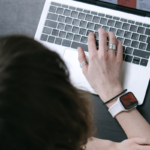 Vista superior de uma pessoa utilizando um notebook sobre uma mesa escura. Uma das mãos está posicionada sobre o teclado e usa anéis nos dedos, além de um relógio inteligente com pulseira clara no pulso. Parte da cabeça e dos braços aparece em primeiro plano. No canto superior esquerdo da imagem há um ícone branco de uma caixa sobre fundo cinza. O ambiente tem iluminação suave e foco nos detalhes do computador e da mão sobre o teclado.