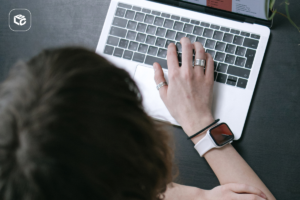 Vista superior de uma pessoa utilizando um notebook sobre uma mesa escura. Uma das mãos está posicionada sobre o teclado e usa anéis nos dedos, além de um relógio inteligente com pulseira clara no pulso. Parte da cabeça e dos braços aparece em primeiro plano. No canto superior esquerdo da imagem há um ícone branco de uma caixa sobre fundo cinza. O ambiente tem iluminação suave e foco nos detalhes do computador e da mão sobre o teclado.